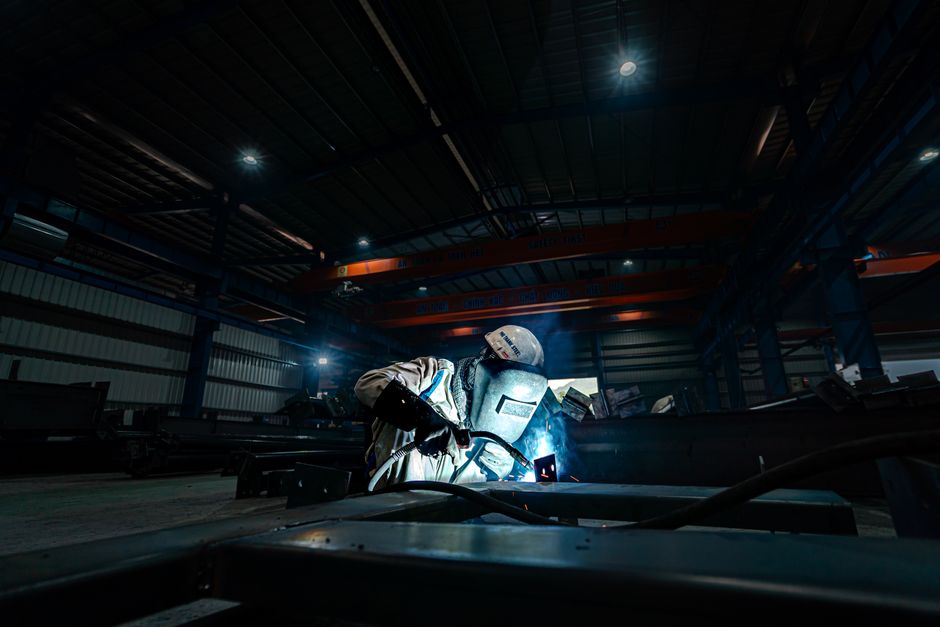 A welder working on steel structures in a large industrial warehouse, emitting bright sparks. (Photo: Hoang NC / Pexels)