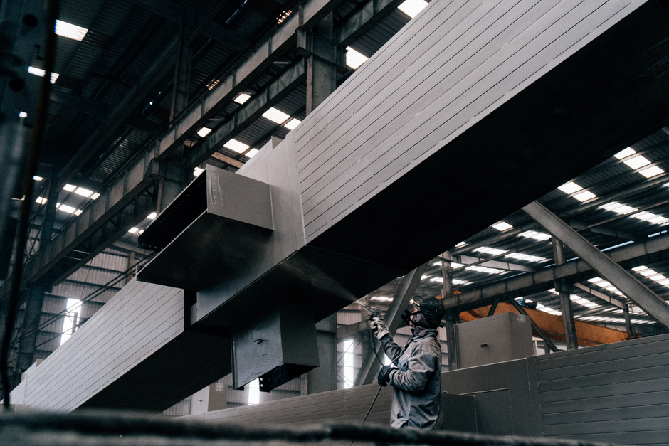 A worker sprays paint on large metal beams in an industrial warehouse. (Photo: Hoang NC / Pexels)