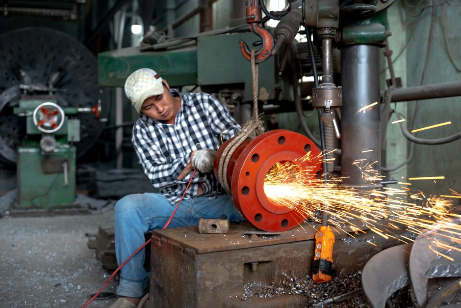 An Asian blue-collar worker operates machinery, grinding metal with sparks flying in a warehouse setting. (Photo: Quang Nguyen Vinh / Pexels)
