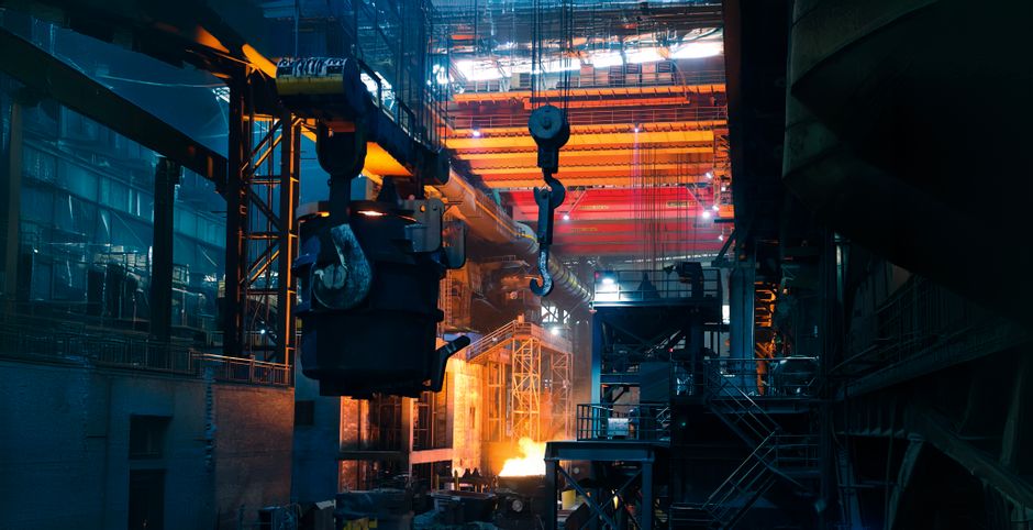 Inside a bustling steel factory with cranes and molten metal casting an intense glow. (Photo: jason hu / Pexels)