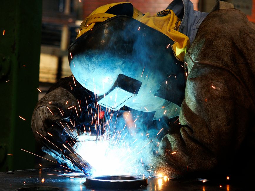 A welder in protective gear creates sparks while working on metal indoors. (Photo: Marcello Sokal / Pexels)