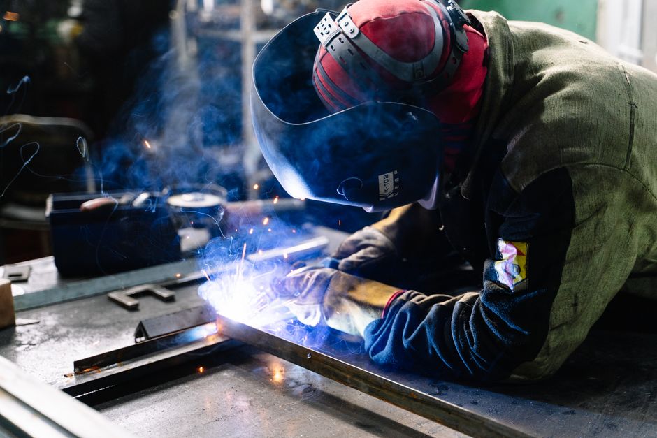 A welder works on metal fabrication indoors, creating bright sparks and smoke. (Photo: Pavel Chernonogov / Pexels)