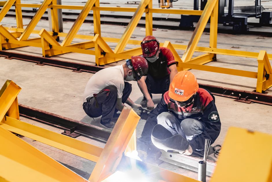 Three construction workers welding metal structures indoors, wearing safety gear. (Photo: Hoang NC / Pexels)