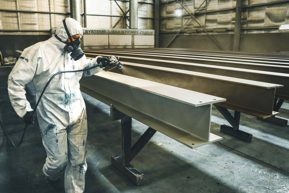 Worker in protective suit spray painting metal beams in an industrial setting indoors. (Photo: Mat Sheard / Pexels)