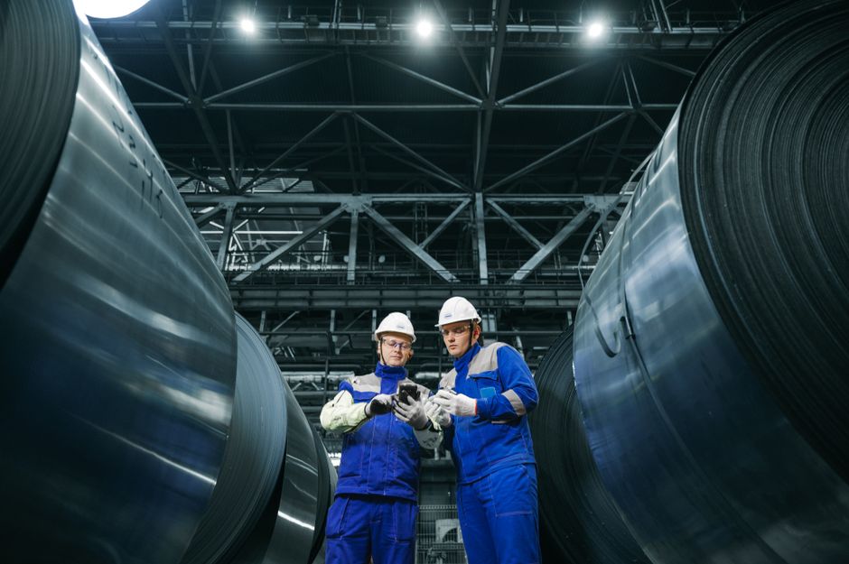 Two workers in protective gear examine steel coils in a modern industrial facility. (Photo: Sergey Sergeev / Pexels)