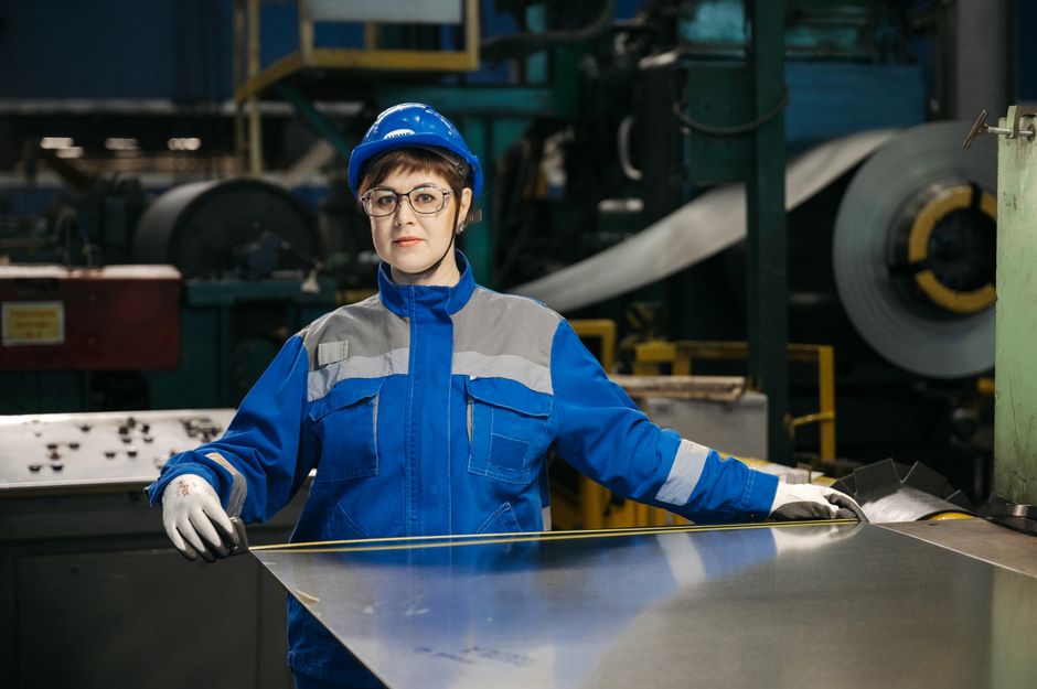 Female factory worker in protective gear handling a metal sheet in an industrial setting. Safety and industry focus. (Photo: Sergey Sergeev / Pexels)