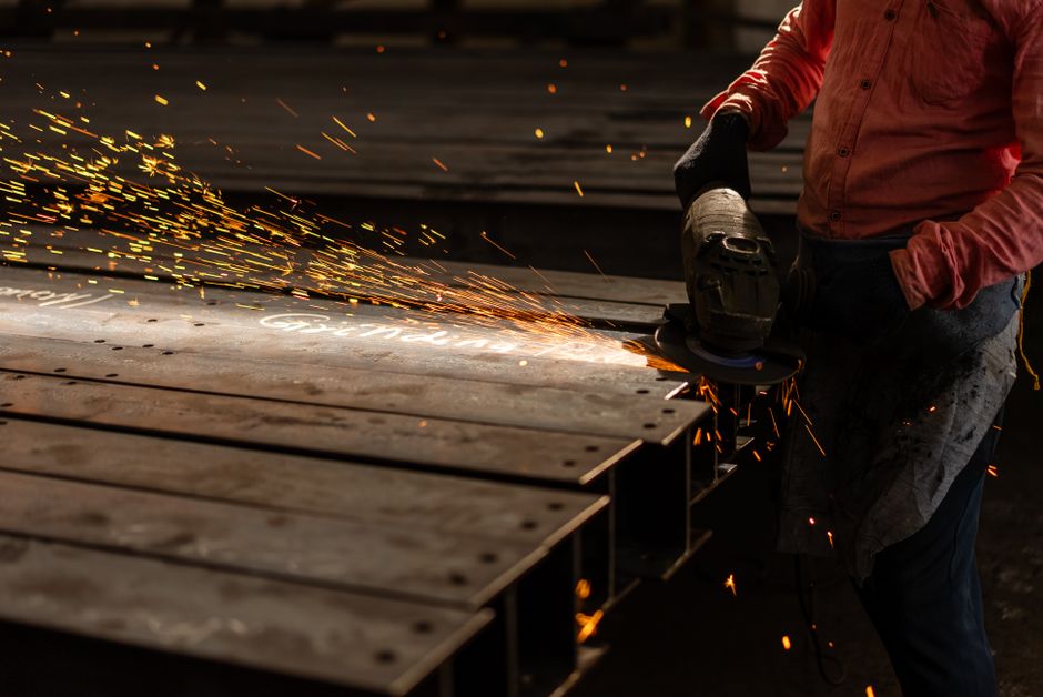 A worker grinds metal beams with an angle grinder, creating vibrant sparks in an industrial setting. (Photo: Michael Orshan / Pexels)